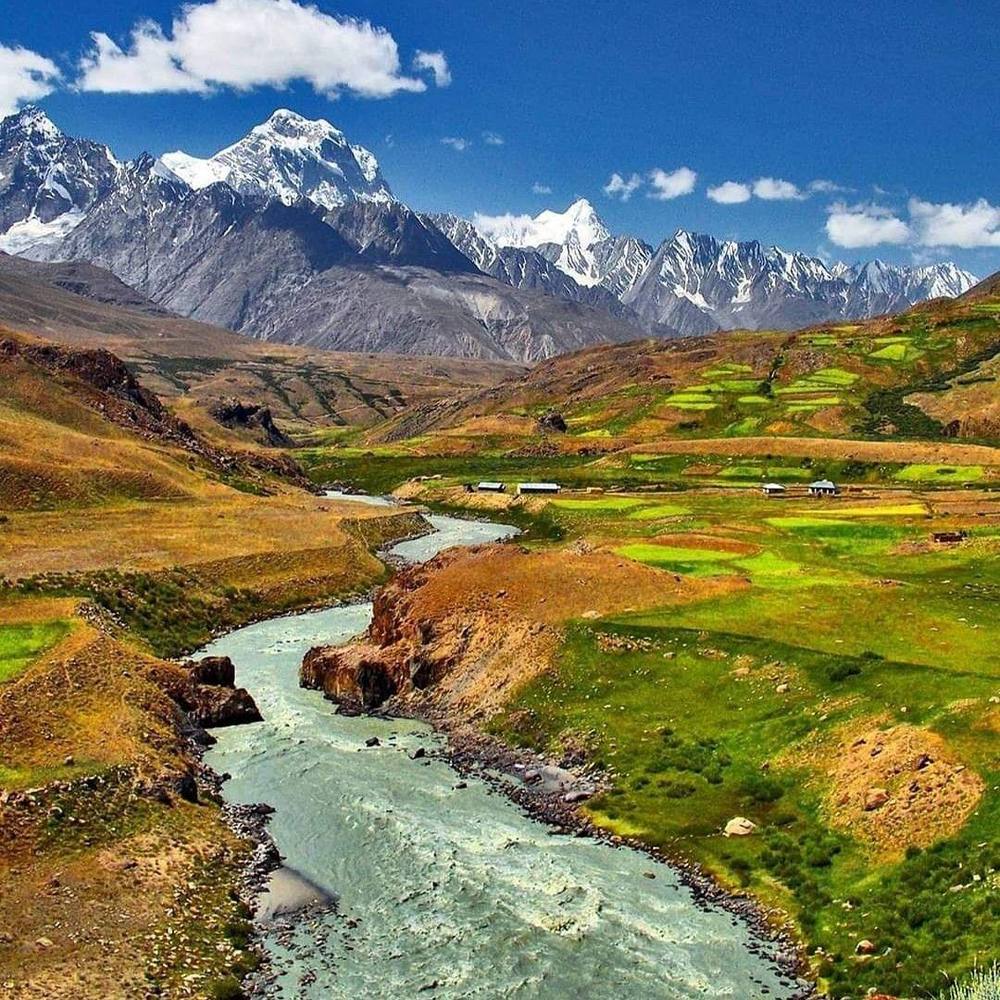 A turquoise river winding through green and golden terraced fields beneath snow-capped Himalayan peaks